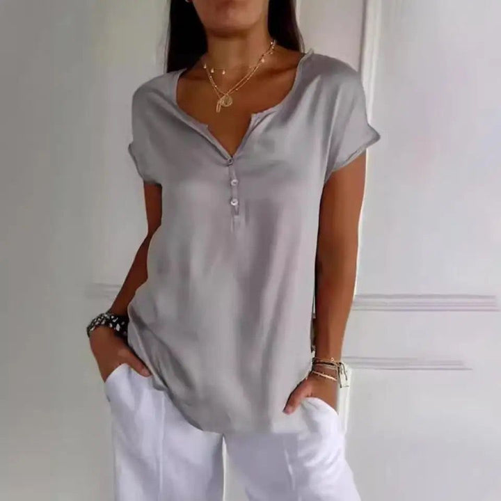 Haut à manches courtes de couleur gris clair avec un décolleté en V et des boutons délicats. Il est porté avec un pantalon blanc ample, créant une tenue moderne, élégante et confortable, idéale pour un style décontracté.