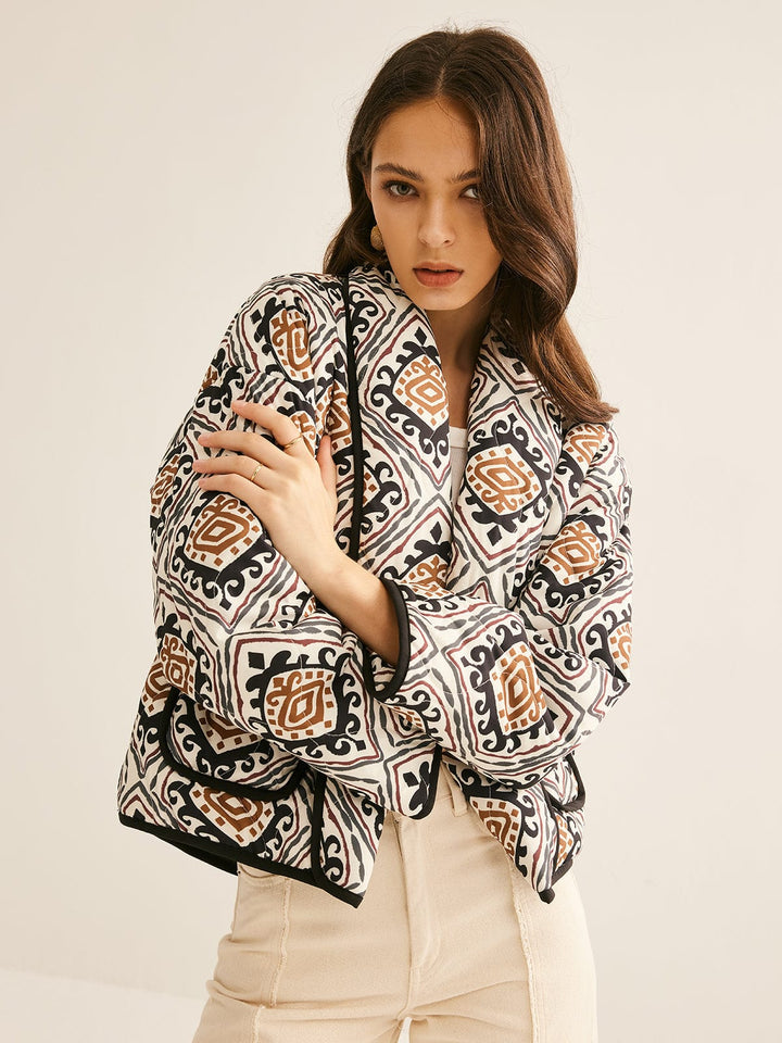 veste ample à motifs géométriques noirs, blancs et bruns, manches longues légèrement bouffantes et bordure noire, portée ouverte sur un haut clair et un pantalon clair dans un cadre neutre, style moderne.