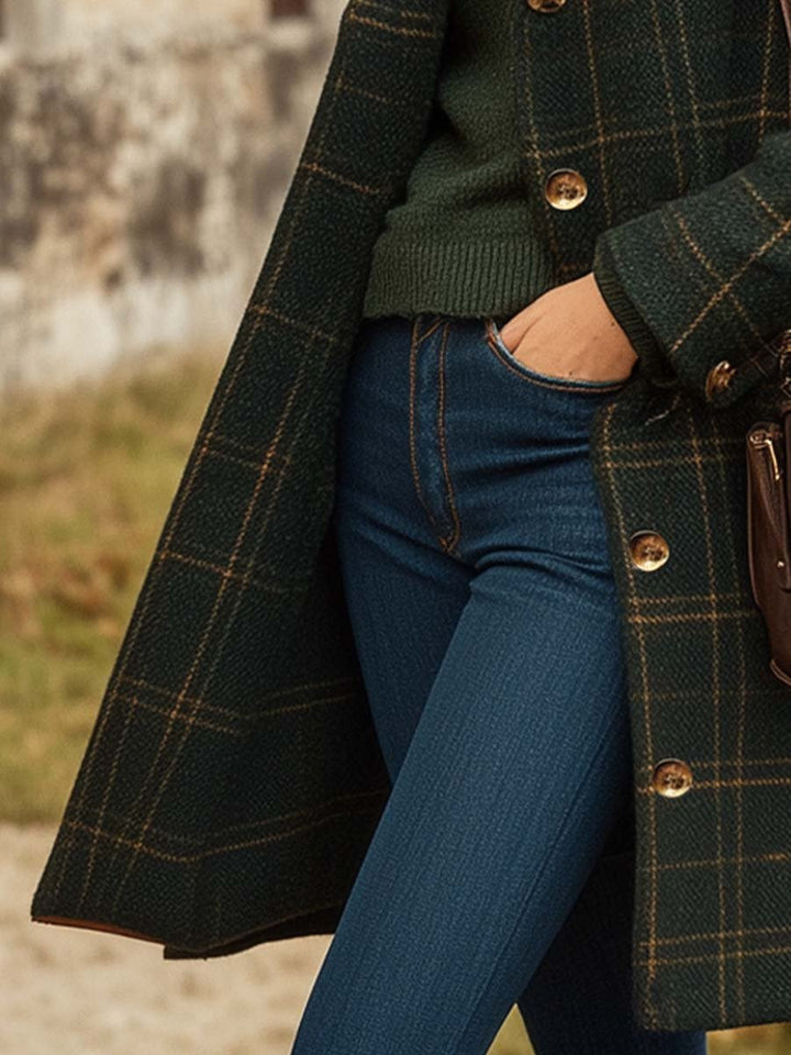Manteau long pour femme à carreaux verts, porté sur un pull chaud et un jean ajusté bleu. Le tissu à motif quadrillé et les boutons dorés apportent une touche élégante et intemporelle. Ce style raffiné est parfait pour affronter la saison froide avec chic et confort.